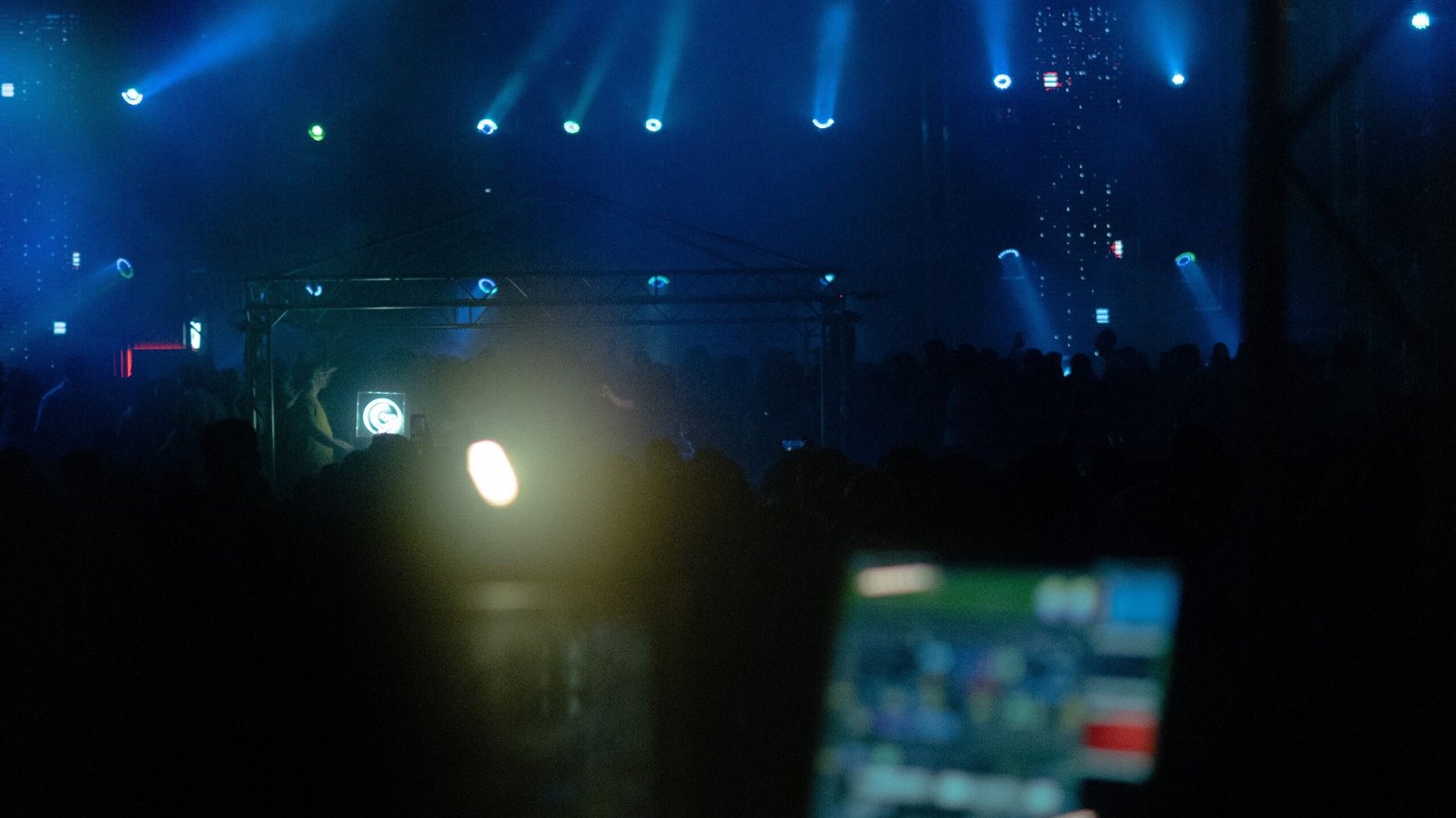 pexels-onbab-13137931 - Sewa Starlink Jakarta Rental Starlink Dark concert venue with blue stage lights piercing through fog and a silhouetted crowd; a laptop screen glows in the foreground.